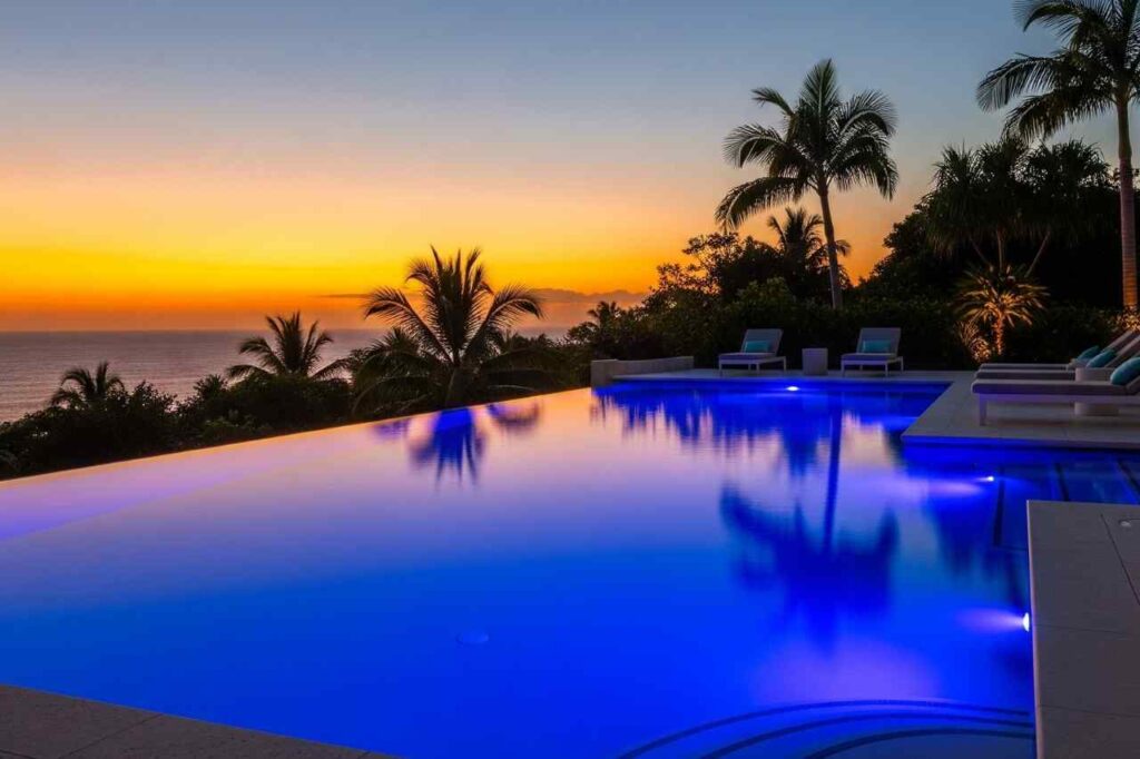 Luxury infinity edge pool in Sydney with blue LED lighting glowing at sunset, surrounded by palm trees and ocean views.